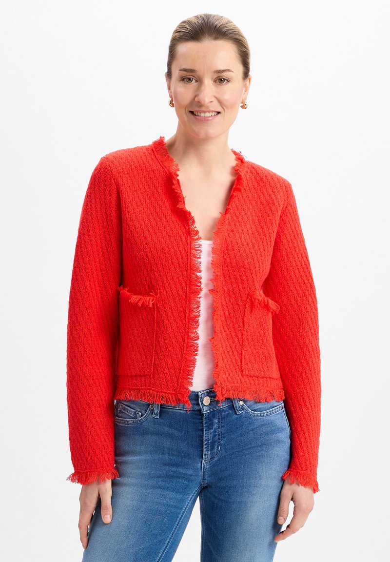 Roter strukturiertes Cardigan mit ausgefransten Kanten, ausgestattet mit zwei Fronttaschen und einem gekürzten Design. Getragen über einem weißen Oberteil und kombiniert mit blauen Jeans.