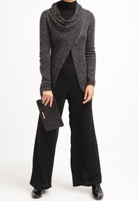 Grå cowl-neck tröja med omslag, långa ärmar och dragkedjedetalj, kombinerad med vida svarta byxor och en svart kuvertväska.