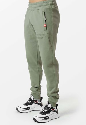 Groene joggerbroek met ritszakken aan de zijkant, gedragen met witte en zwarte sportschoenen op een witte achtergrond.