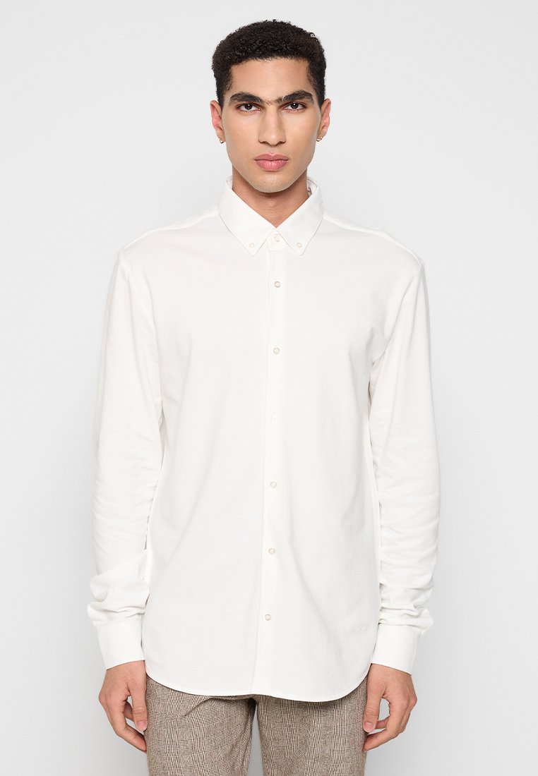 Młody mężczyzna z krótkimi, kręconymi włosami, ubrany w białą koszulę z długim rękawem zapinaną na guziki i jasnobrązowe spodni w kratkę, stojący na tle jednolitego tła.