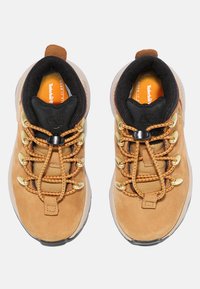 Chaussures de randonnée en suede beige avec des cols rembourrés noirs, des œillets en métal doré et une semelle en caoutchouc texturé. Présente une marque orange sur la languette.