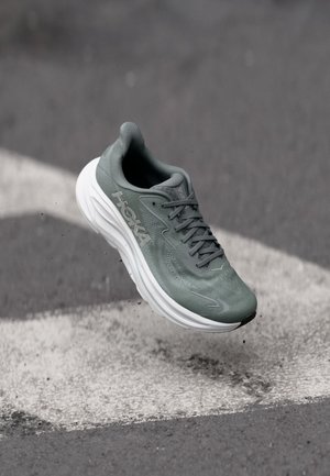 Zaļš Hoka skriešanas apavs ar baltu zoli gaisā virs pelēka un balta bruģa fona ar struktūru.