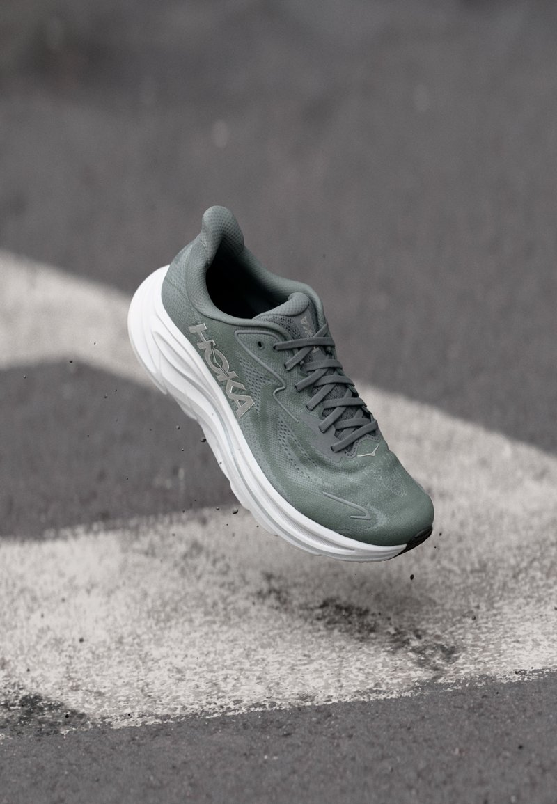 Πράσινο παπούτσι τρεξίματος Hoka με λευκή σόλα, στον αέρα πάνω από υφή γκρι και λευκής πλακόστρωτης επιφάνειας.