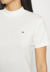 Bílý krátký rukáv tričko s vysokým límcem, na kterém je malé logo v modré, červené a bílé barvě. Vyrobeno z měkké bavlny.