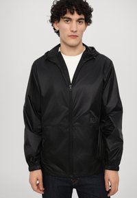 Veste légère noire avec capuche, fermeture éclair à l'avant, poignets élastiques et logo EA7. Texture lisse, design minimaliste et coupe décontractée.