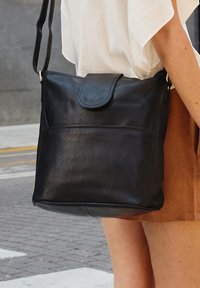 Melna ādas plecu soma ar vāku, gluda tekstūra, priekšējais kabatiņa un gara regulējama siksna. Kvadrātveida forma un minimāls dizains.