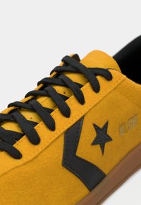 Sapatilha de camurça amarela com atacadores pretos, logotipo de estrela preta e chevron, e o texto "ALL STAR" na lateral, com uma sola castanha.