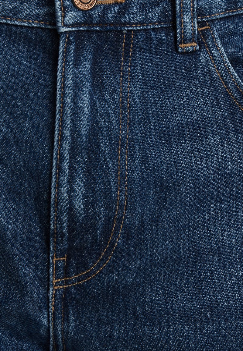 Gros plan sur un jean en denim bleu présentant la poche avant, la passant de ceinture, le bouton et des détails de couture orange.
