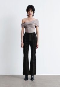 Beige Oberteil mit schulterfreiem Design und drapierter Stoff, kombiniert mit hochgeschnittenen schwarzen Schlagjeans mit kontrastierenden Nähten und stylischen schwarzen Stiefeln.