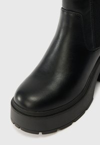 Botín de cuero negro hasta el tobillo con puntera redondeada, panel lateral elástico texturizado y suela de goma negra gruesa con detalles de superficie sutiles.