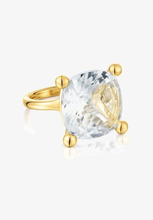 Anello d'oro con grande pietra preziosa trasparente taglio cuscino, sostenuta da quattro castoni arrotondati in oro, mostrato su superficie bianca riflettente.