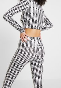 Žena oblečená v černobílém pruhovaném crop topu s logem Adidas a těsných legínách, stojí s rukama na bocích.