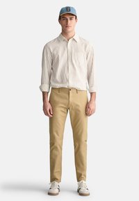 Lysstribet skjorte med knapper, khaki bukser og en blå cap med logo; iført hvide sneakers med sorte detaljer og beige sål.