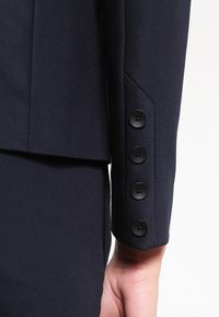 Marinblå kavajsärm med slät tyg, skarp skärning och fyra svarta knappar vid manschetten. Skräddarsydd design med rena linjer.