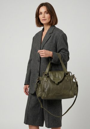 Woman in gray blazer and skirt holding olive green leather handbag with flap and multiple straps against plain background.