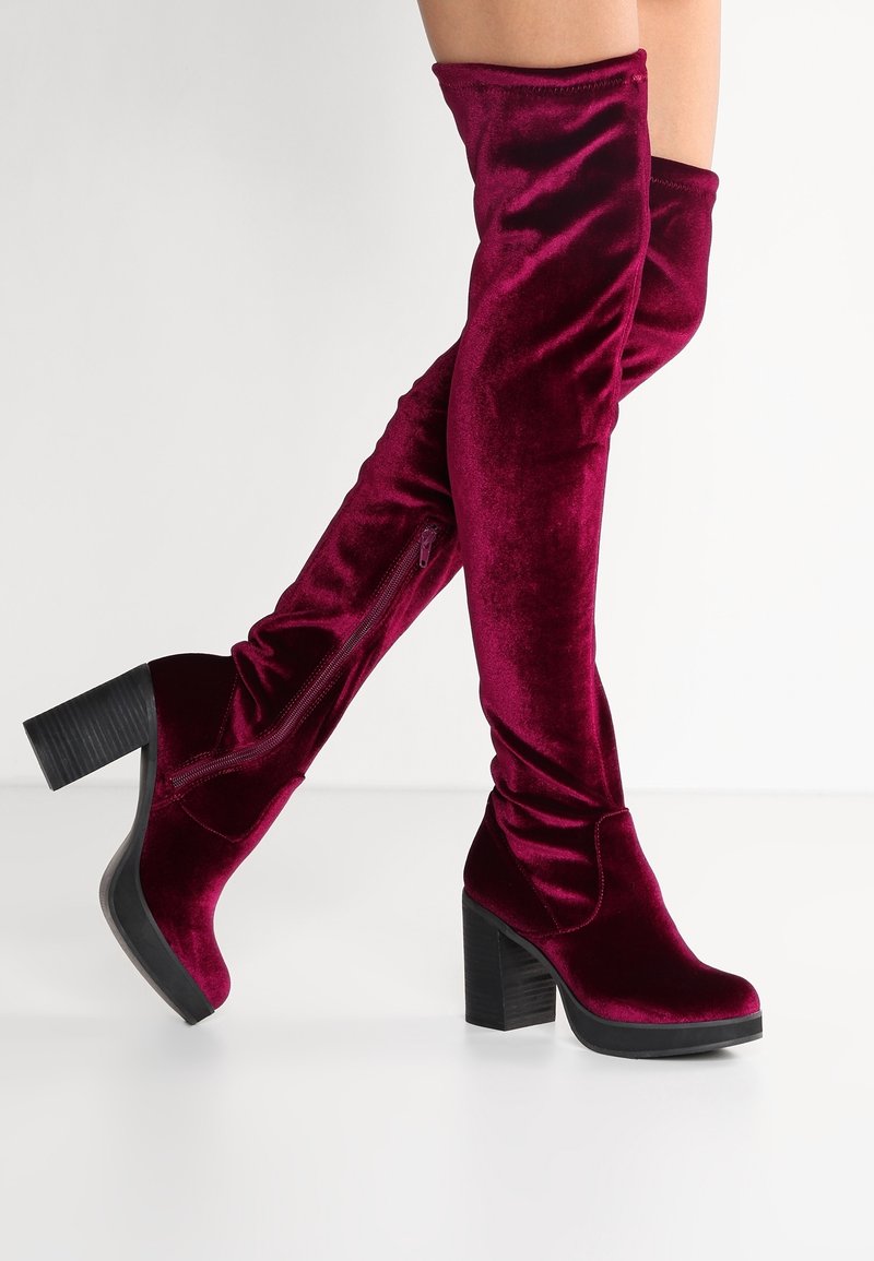 Bottes en velours pour femmes, hauteur genou, de couleur bordeaux foncé, avec talons blocs et fermetures éclair latérales, portées sur des jambes croisées sur fond blanc.
