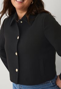 Black cropped jacket with a pointed collar, gold buttons, and front pocket detail. Smooth texture, tailored fit, suitable for layering.