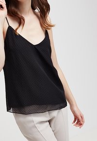 Zwarte doorschijnende cami met spaghettibandjes, voorzien van een stippenpatroon en een losse pasvorm, gecombineerd met lichte broeken.