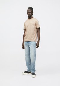 Lichtbruin katoenen T-shirt met een zakje en een klein logo, gecombineerd met verweerde blauwe spijkerbroek en zwarte sneakers.