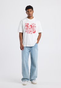 White t-shirt with red graphic design featuring abstract figures. Paired with light blue wide-leg jeans and white sneakers. Casual look.