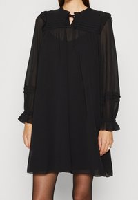 Vestido negro de manga larga con escote plisado, detalle de ojal y puños con volantes. Confeccionado en una tela ligera y transparente, con una silueta en línea A que fluye.