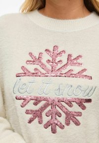 Baltas megztinis su rožinės spalvos blizgančiu snaigės raštu ir išsiuvinėtu užrašu „let it snow“, minkštu, tekstūruotu paviršiumi.
