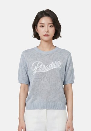 Jonge vrouw met kort donker haar die een lichtblauwe gebreide trui draagt met "Paradise" erop geschreven en een witte broek, staand tegen een effen achtergrond.