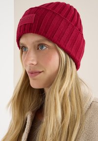 Rode geribbelde gebreide beanie met een textuurafwerking en een rechthoekig stoffen label aan de voorkant. Zacht materiaal met een aansluitend ontwerp.