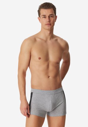 Oberkörperfreier Mann, der steht und graue Boxershorts mit schwarzen Seitenstreifen vor einem schlichten weißen Hintergrund trägt.