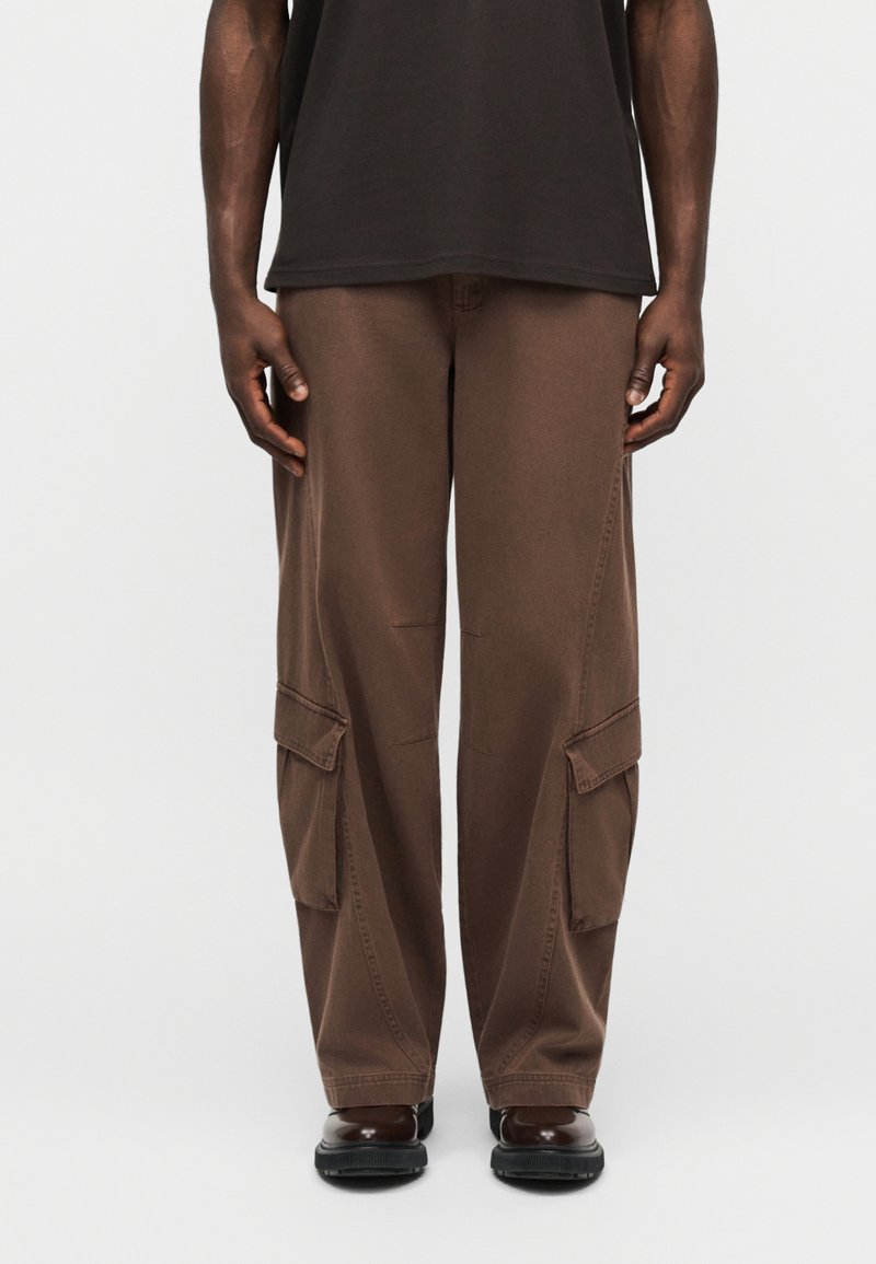 Άτομο που φοράει φαρδιά καφέ παντελόνια cargo με μεγάλες πλαϊνές τσέπες, σκούρα καφέ παπούτσια και μαύρο πουκάμισο, στέκεται μπροστά σε απλό φωτεινό φόντο.