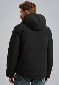 Giacca isolante nera con cappuccio, caratterizzata da una texture liscia e un design minimalista. Cuciture visibili e dettaglio sui polsini delle maniche.