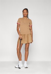 Beige golfoutfit bestående av en texturerad pikétröja med rosa detaljer och en plisserad kjol. Modellen håller en golfklubba och bär vita sneakers.