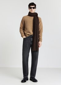 Mann mit Sonnenbrille, braunem Pullover, langem dunkelbraunem Schal, schwarzen Jeans und schwarzen Stiefeln, der mit einer Hand in der Tasche vor einem weißen Hintergrund steht.
