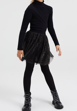 Niño con camiseta negra de manga larga acanalada, falda plisada brillante, medias negras y botines negros con cordones, de pie sobre fondo blanco.