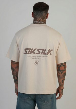 Mand med tatoveret nakke og arme bærer beige oversized T-shirt med "SikSilk" og slogan trykt på bagsiden, kombineret med blå jeans.