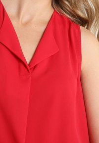 Punainen, hihaton paita, jossa on kauluksellinen pääntie ja pehmeä, laskeutuva kangas. Malli on sileä ja siinä on vähän ompeleita.