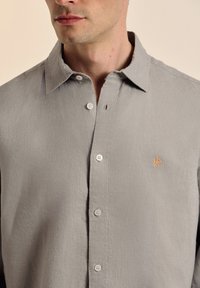 Camisa de botones en gris claro, hecha de una tela suave. Presenta un cuello con punta, tapeta delantera con botones y un pequeño logotipo naranja encima del bolsillo izquierdo del pecho.