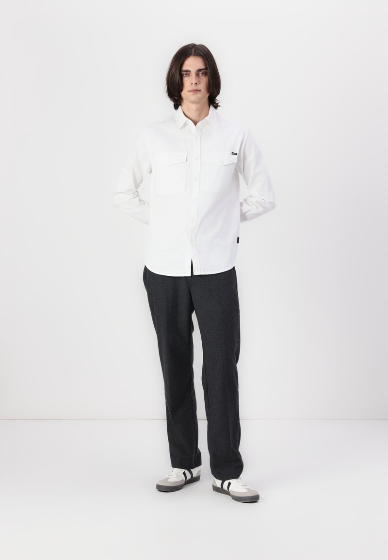 Jonge persoon met schouderlang donker haar staat met de handen achter de rug, gekleed in een witte blouse, donkere broek en sneakers.