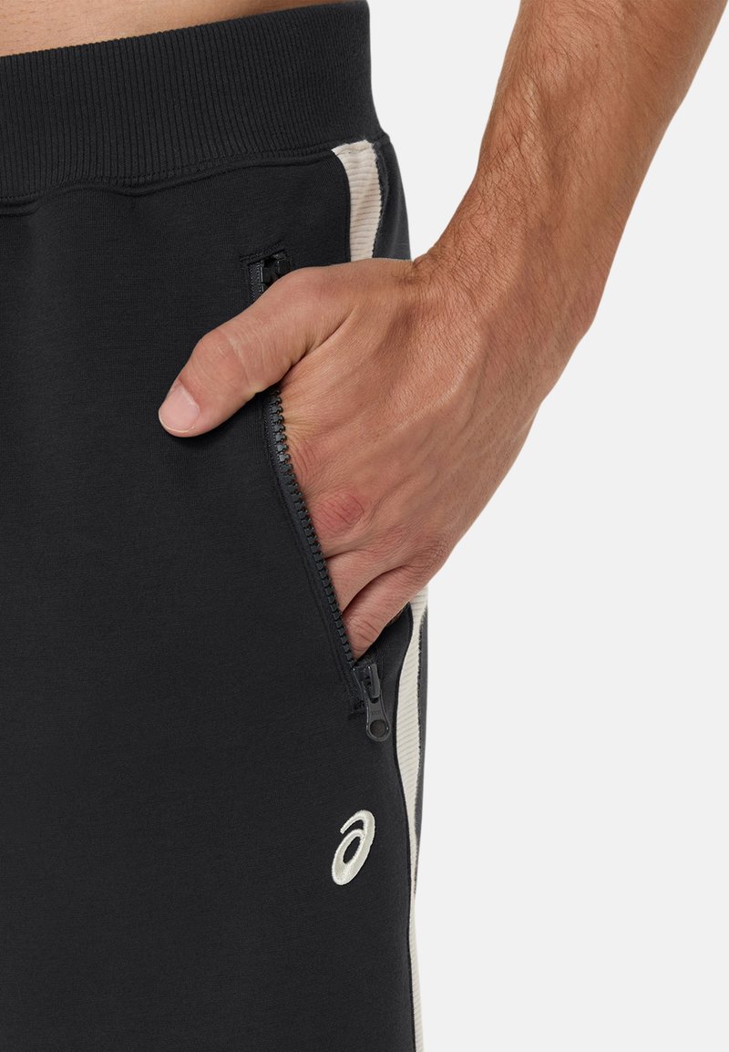 Jogging noir avec poche zippée, dotée d'une bande blanche sur le côté. Texture douce avec une ceinture élastique et un détail de logo brodé.