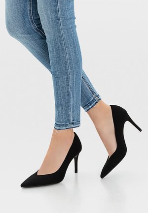 Personne portant un jean skinny bleu clair et des escarpins noirs à bouts pointus, se tenant avec un pied légèrement levé sur un sol blanc.