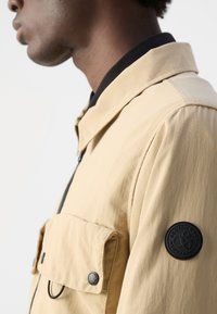 Beige lichtgewicht jas met een ritssluiting, twee borstzakken en een zwarte patch op de mouw. Soepele textuur en casual kraagontwerp.