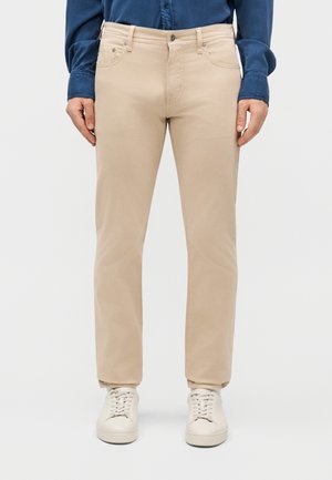 Άνδρας που φοράει μπεζ παντελόνι ίσιας γραμμής, λευκά αθλητικά παπούτσια και σκούρο μπλε μακρυμάνικο πουκάμισο, στέκεται μπροστά σε απλό φόντο.