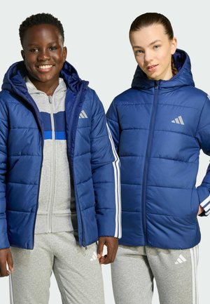Zwei lächelnde junge Menschen tragen blaue Daunenjacken über grauer Sportbekleidung und posieren vor einem hellen Hintergrund.
