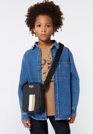 Borsa a tracolla nera con strisce orizzontali beige e bianche, realizzata in materiale sintetico. Indossata da un bambino con capelli ricci, in una giacca di jeans.