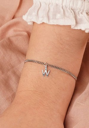 Silbernes Kettenarmband mit einem hängenden "W"-Anhänger am Handgelenk, begleitet von einem weißen strukturierten Ärmel und einem weichen rosa Stoffhintergrund.