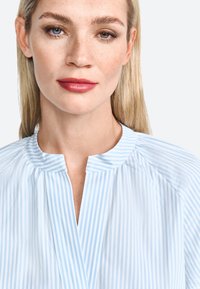 Blusa a righe di colore azzurro chiaro con colletto alla mandarino, caratterizzata da righe verticali e una patta senza bottoni. Texture liscia con una leggera lucentezza.