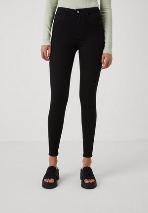 Femme portant un jean skinny noir taille haute, un haut vert clair à manches longues côtelé, et des sandales à plateforme noires à bout ouvert, debout sur un fond uni.