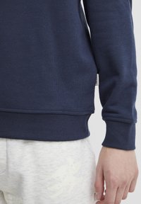 Navy blue sweatshirt featuring a ribbed cuff and hem, made of soft cotton blend material; displayed alongside light grey pants with subtle design.