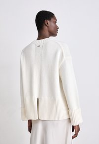 White knitted sweater with ribbed cuffs and hem, featuring a back slit and a subtle logo detail at the neckline. Smooth texture.