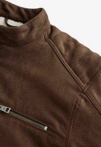 Giacca in suede marrone con colletto alto, dettagli di cucitura sottili e una tasca con zip sul davanti. Texture morbida con finitura opaca.